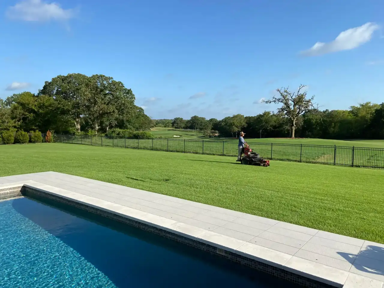 Crew member mowing a large backyard lawn with a stand-on mower beside a pool